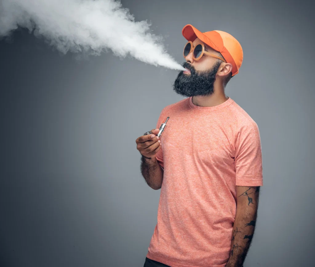 Un homme tatoué barbu joyeux fume une cigarette électronique isolée sur fond gris. 